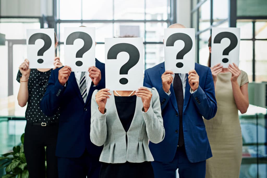 An array of business professionals stand shoulder to shoulder, each obscuring their own face with a placard displaying a question mark