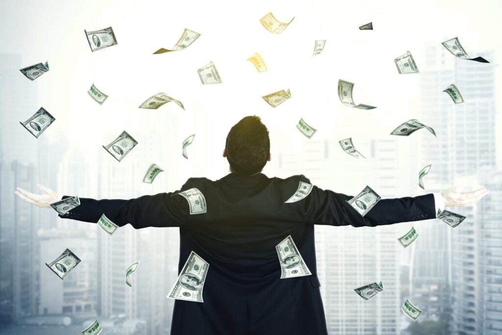 A young entrepreneur holds their arms out wide to welcome one hundred dollar bills raining down from the sky in the business district of a large city.