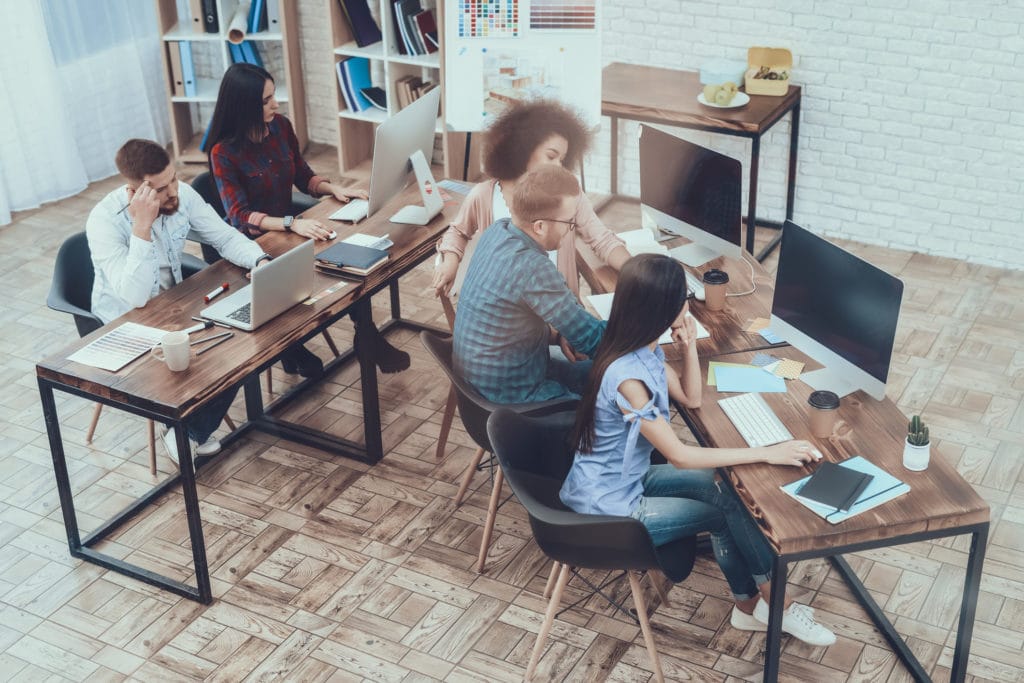 A group of young professionals collaborate in an work space furnished with large table desks, white brick and wood tiles.